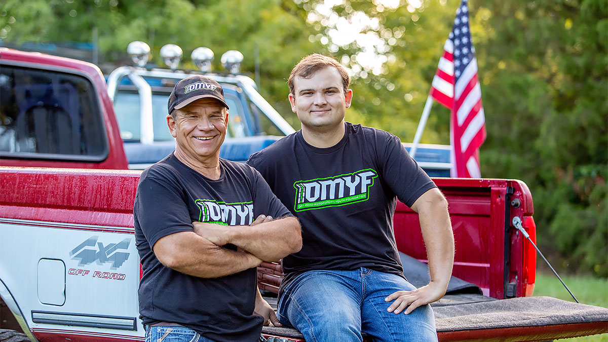 Preston Lewis and his father wearing Outdoor Motorsports Youth Foundation shirts stand near pickup trucks outdoors