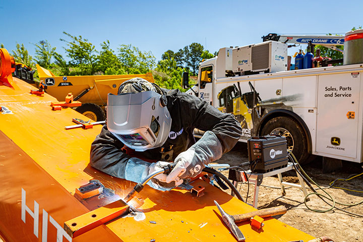 Service technician welds on a piece of heavy equipment in the field with his work truck in the background