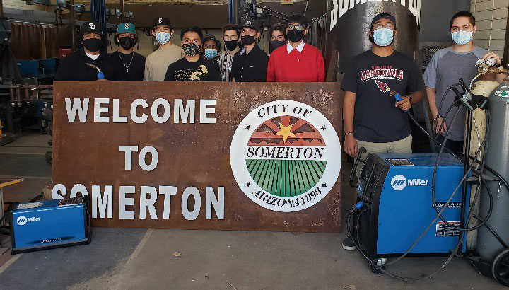 Welding class posing with Welcome to Somerton sign and Miller welders