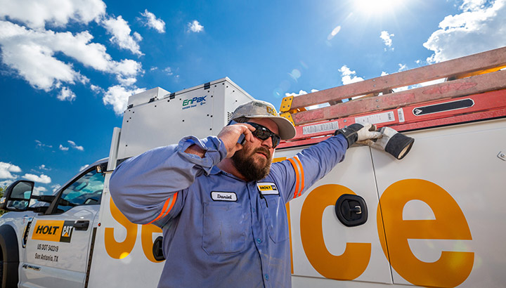 A service truck technician stands next to his truck talking on the phone with the bright sun behind him