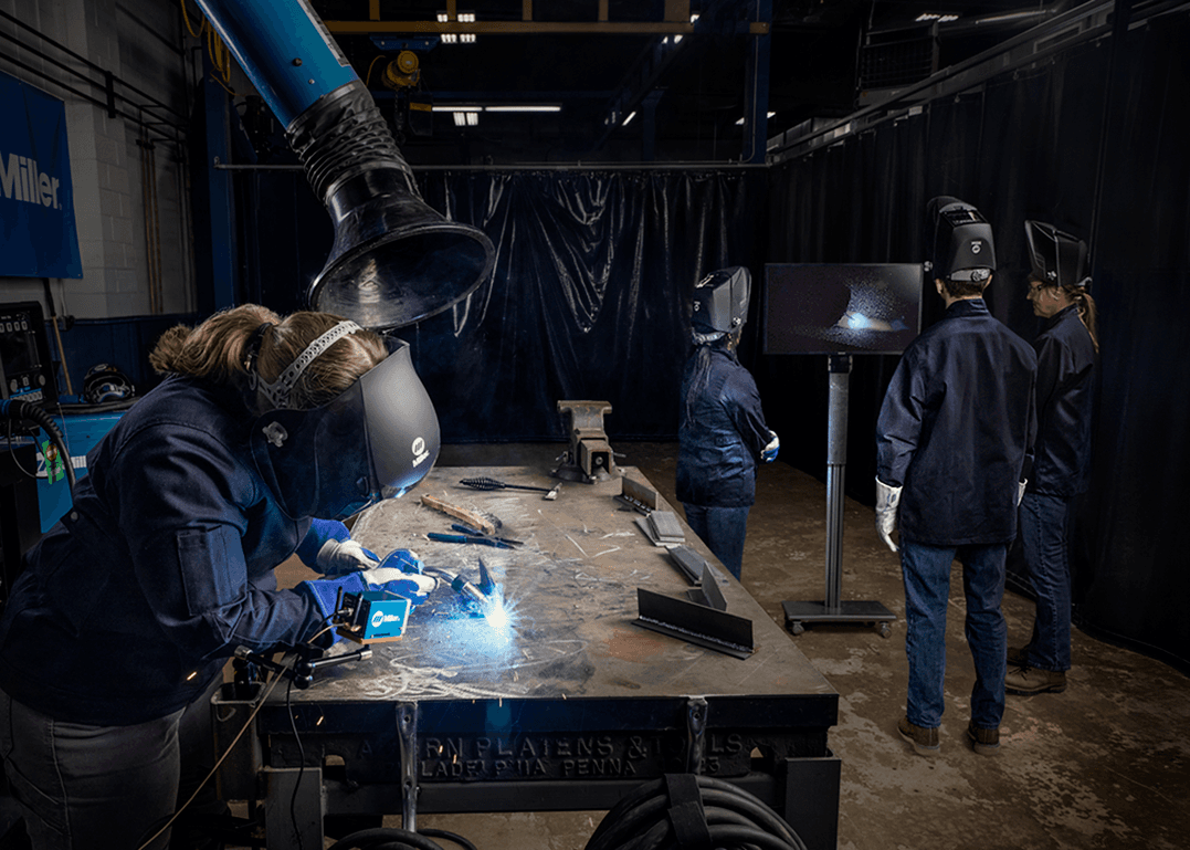 A group of people in welding gear working at a table in a workshop; one person welds while others observe a monitor displaying welding activity.