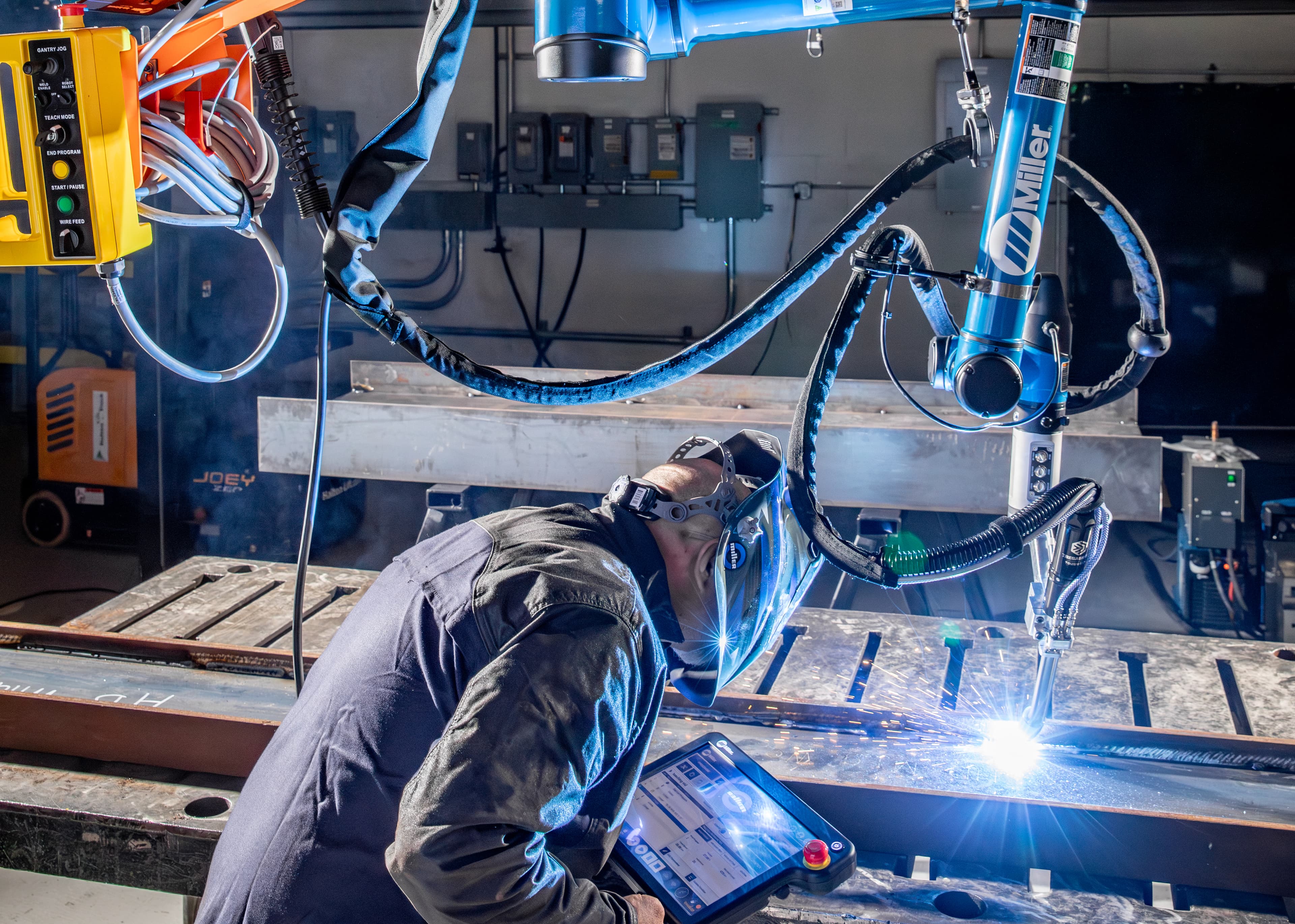 Worker operating a robotic welding arm in a manufacturing facility, with bright sparks and industrial equipment visible.