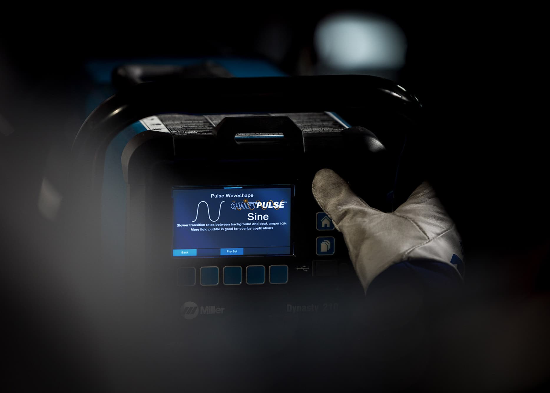 A gloved hand operates the control panel of a Miller Dynasty 210 welding machine, displaying settings for pulse waveform and mode.