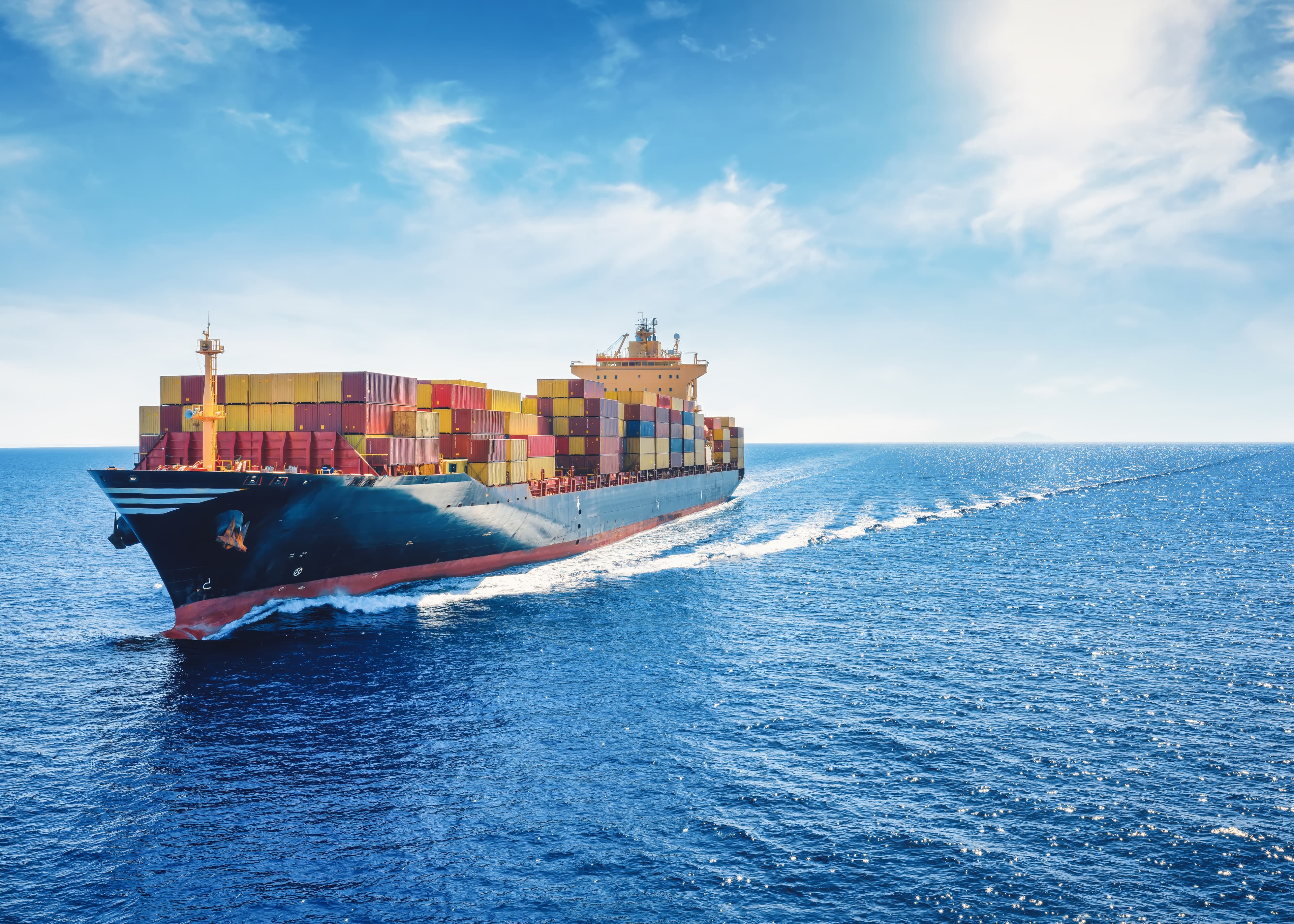A cargo ship carrying numerous colorful containers navigates through the ocean under a bright blue sky.