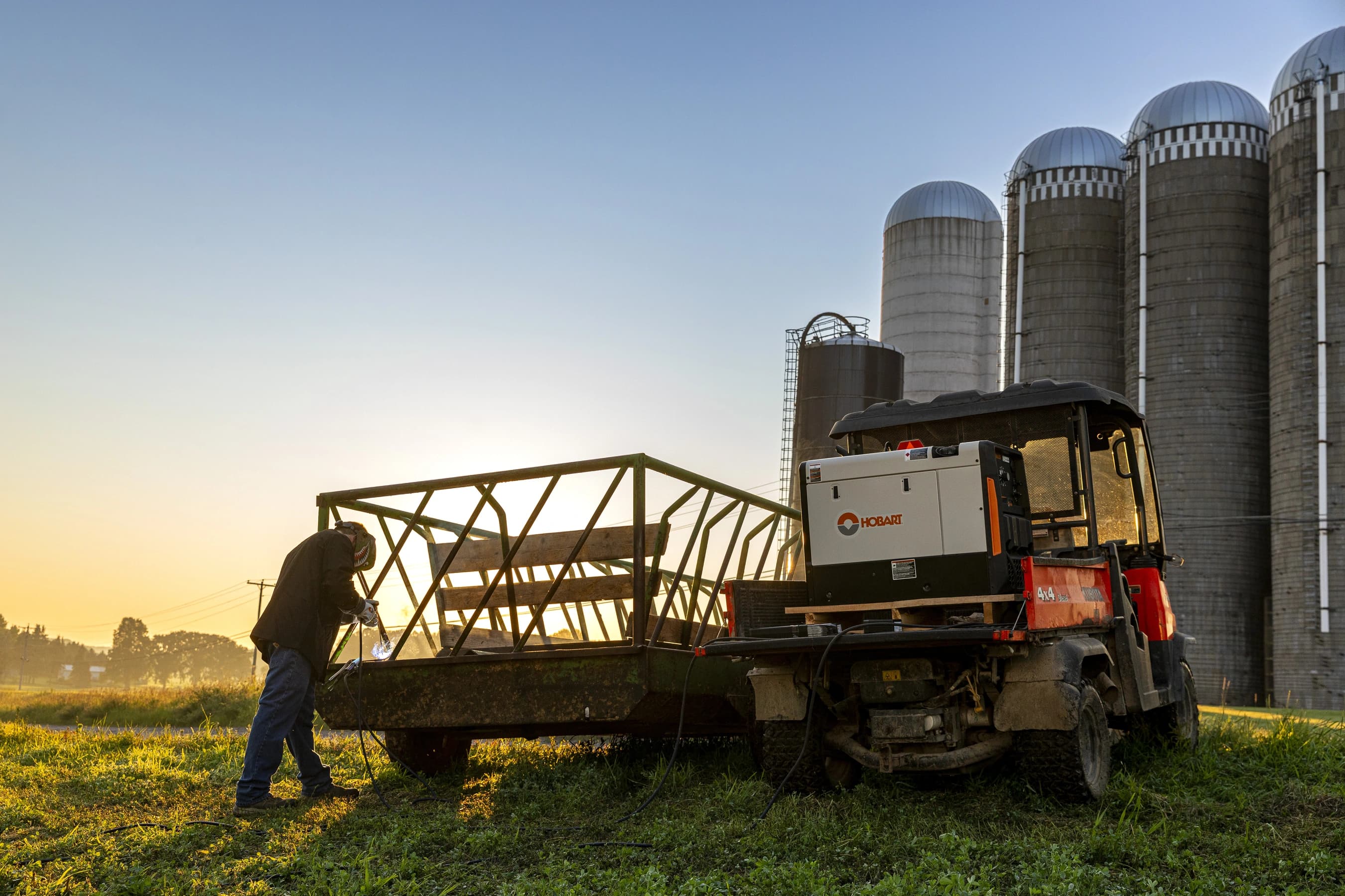 Champion 225 Larrand Dairy Application mml12156 A worker performing welding on a metal structure near farming silos during sunrise. A utility vehicle with Hobart equipment is nearby.