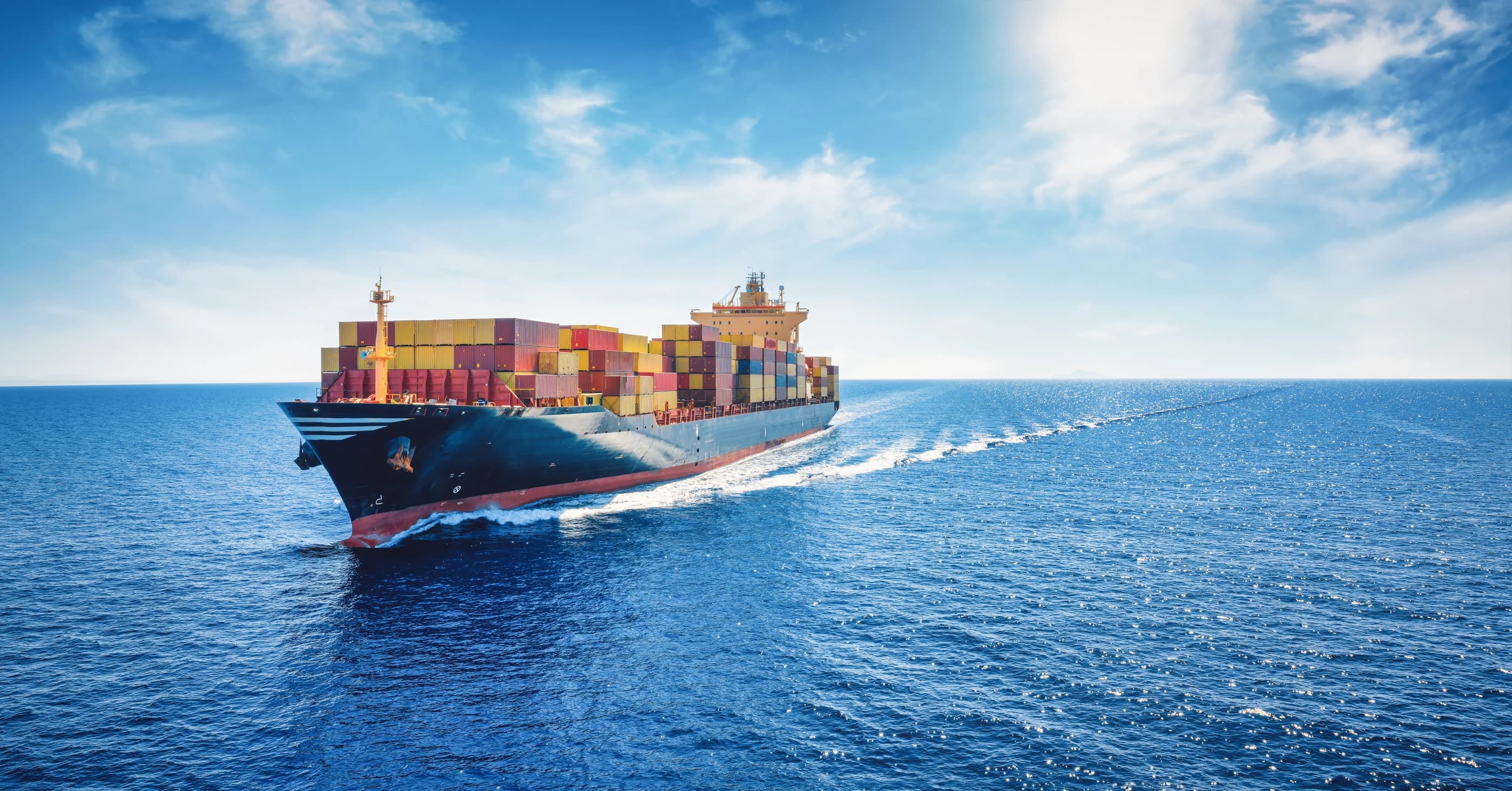 A cargo ship carrying numerous colorful containers navigates through the ocean under a bright blue sky.
