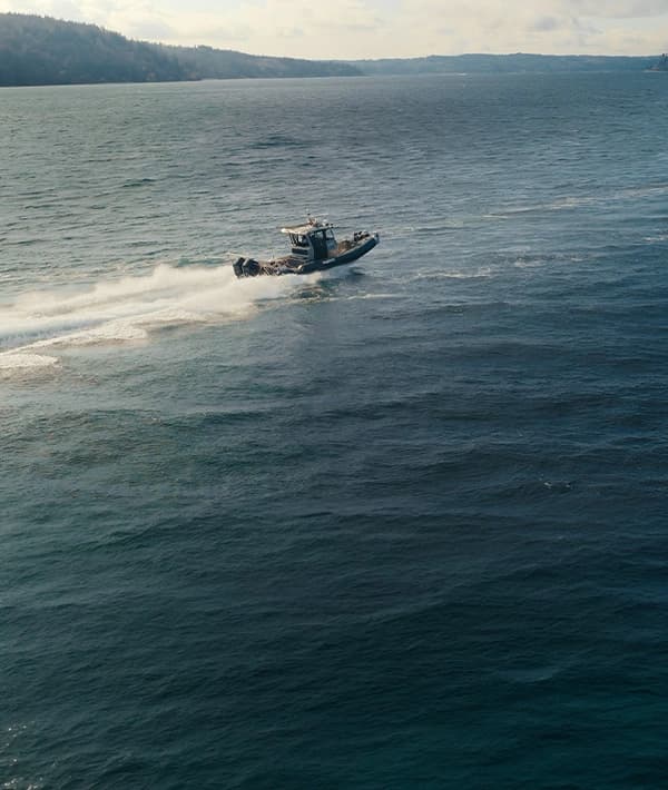 A small speedboat traveling quickly across a large body of water under a cloudy sky, with distant forested shorelines visible.