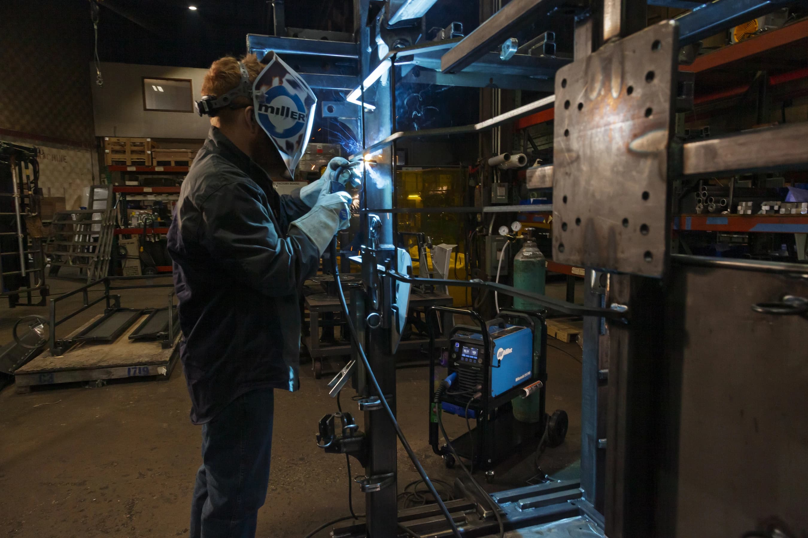 A person welding in a workshop with industrial equipment, wearing protective gear including gloves and a helmet.